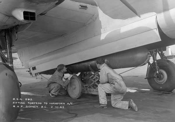 Photographie en noir et blanc – Deux grosses roues visibles sous un avion, un compartiment ouvert et deux hommes accroupis en train de charger une énorme bombe (de 6 à 8 pieds). La bombe est placée sur un cric.